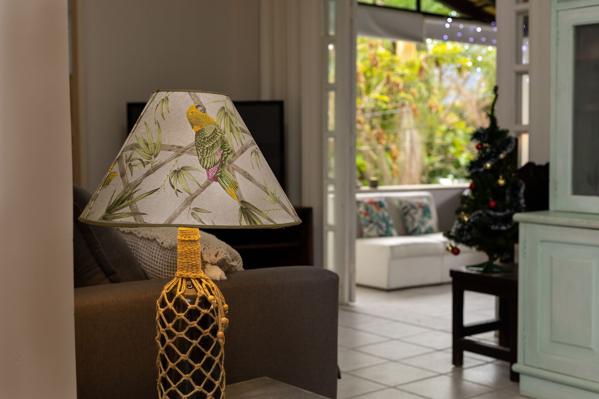 A lamp with a bird design on its lampshade, a television on a stand, a sofa, a Christmas tree, and a view of greenery outside.