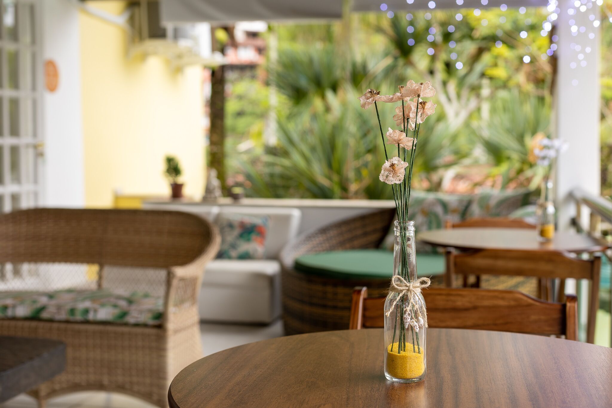 A table with a glass vase containing dried flowers and sand, set on a wooden table in a cozy outdoor seating area with wicker furniture and a green cushion.