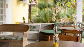 A table with a glass vase containing dried flowers and sand, set on a wooden table in a cozy outdoor seating area with wicker furniture and a green cushion.