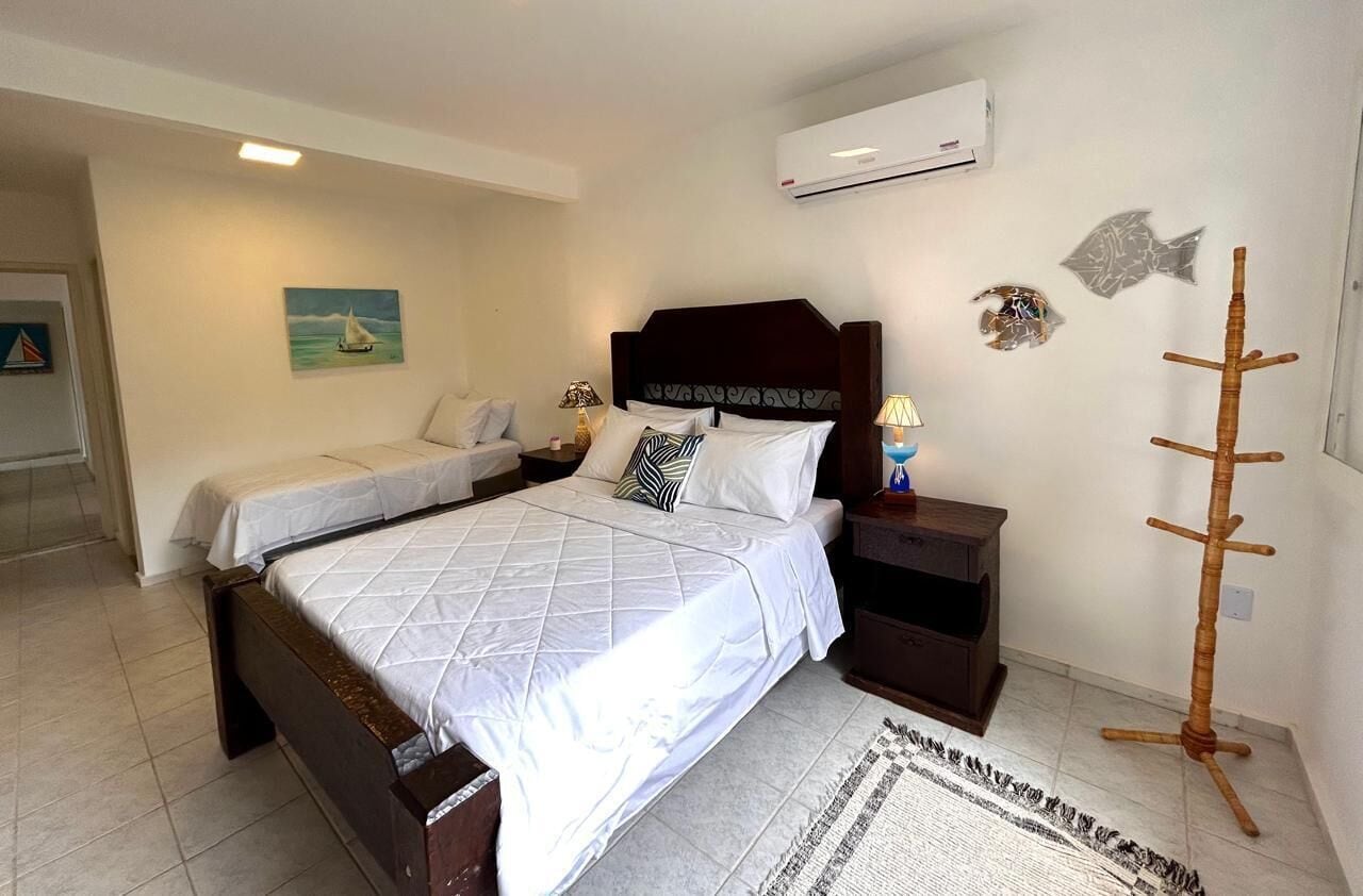 A double bed with a wooden headboard, a bedside table with a lamp, and a small shelf. There is a wall-mounted air conditioner and decorative fish on the wall.
