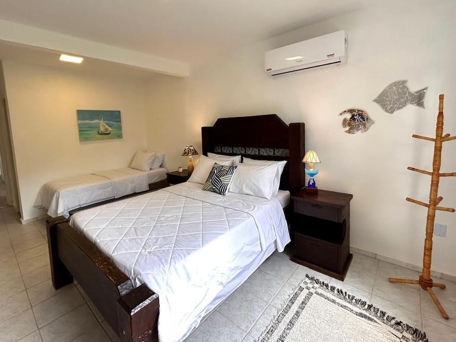 Una cama doble con cabecera de madera, una mesita de noche con lámpara y un estante pequeño. Hay un aire acondicionado de pared y peces decorativos en la pared.