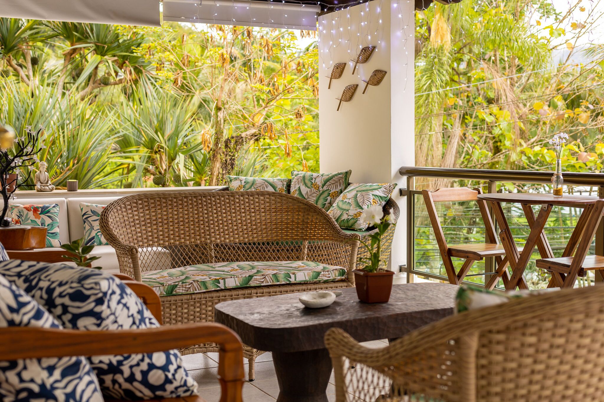 A wicker sofa with a leaf-patterned cushion, a small table with a bowl, and chairs on a balcony with a view of lush greenery.