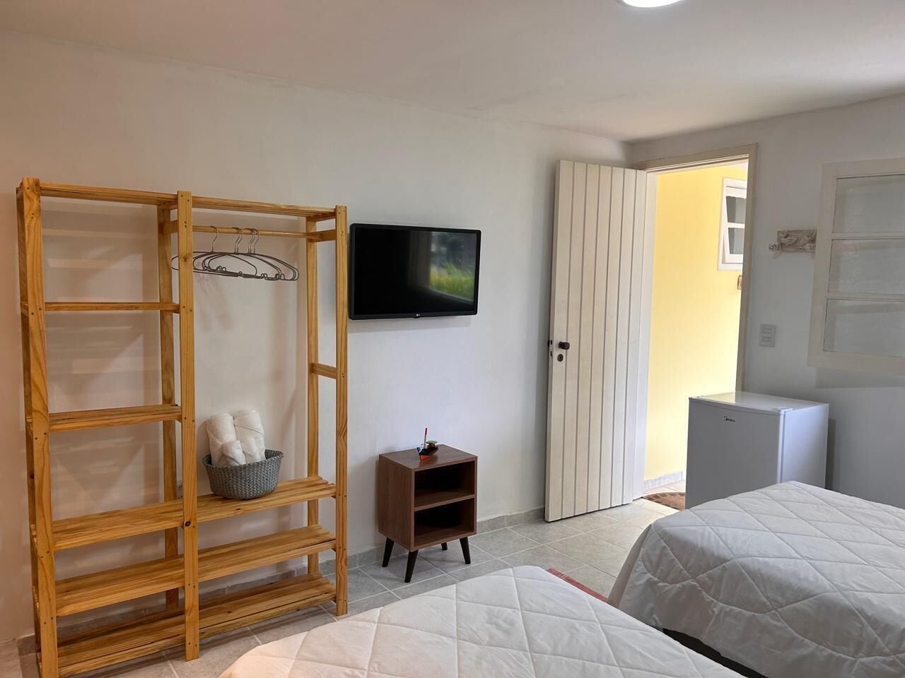 Un dormitorio con un armario de madera, un televisor montado en la pared, una mesita de noche pequeña y una cama con colcha blanca.