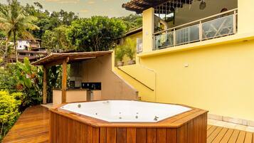 Een houten jacuzzi op een terras met een geel gebouw op de achtergrond.