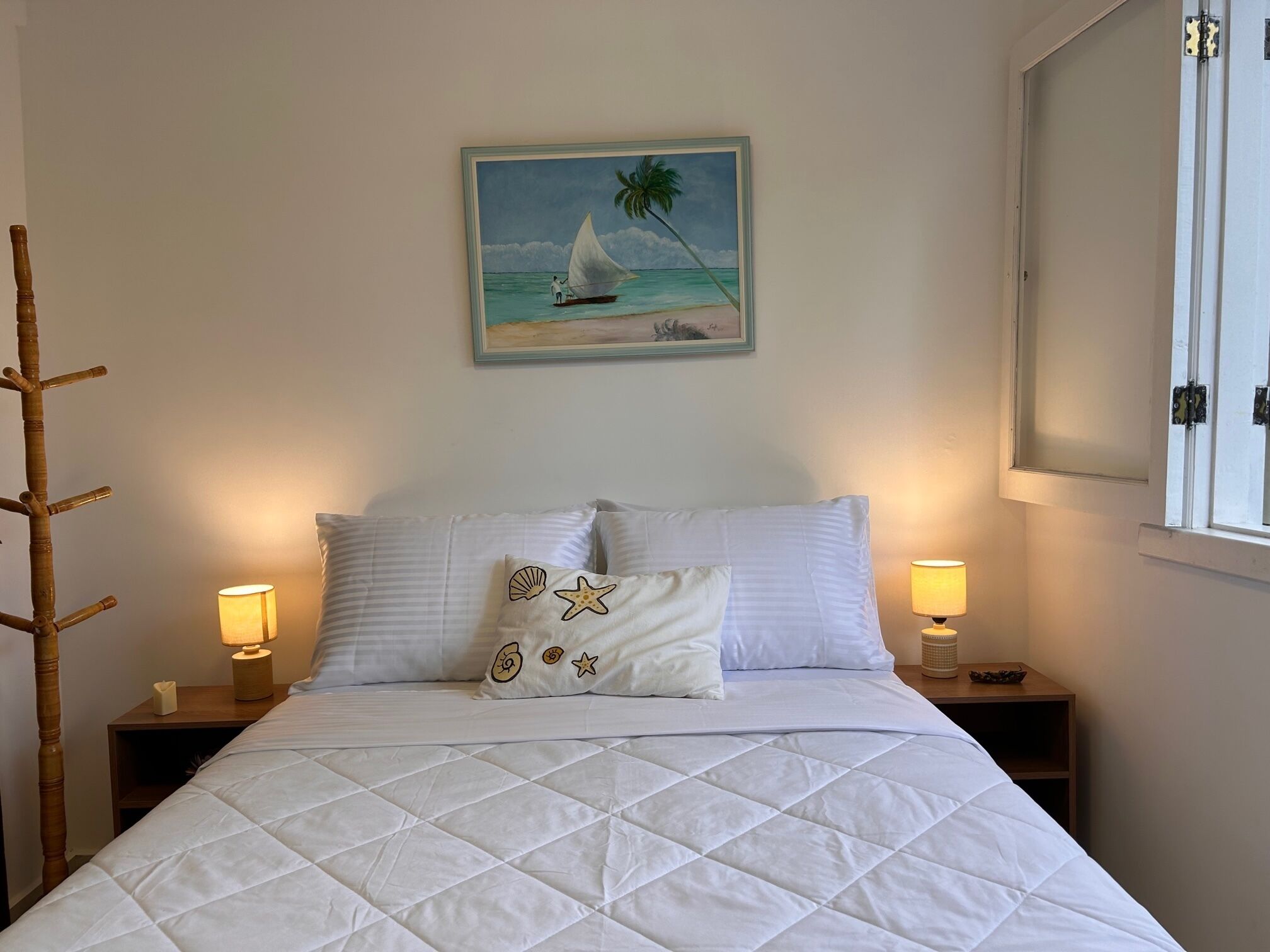 Un lit bien fait, recouvert d’une couette blanche matelassée, deux lampes de chevet, une table de nuit en bois, un tableau représentant un voilier accroché au mur, et une fenêtre à cadre blanc.