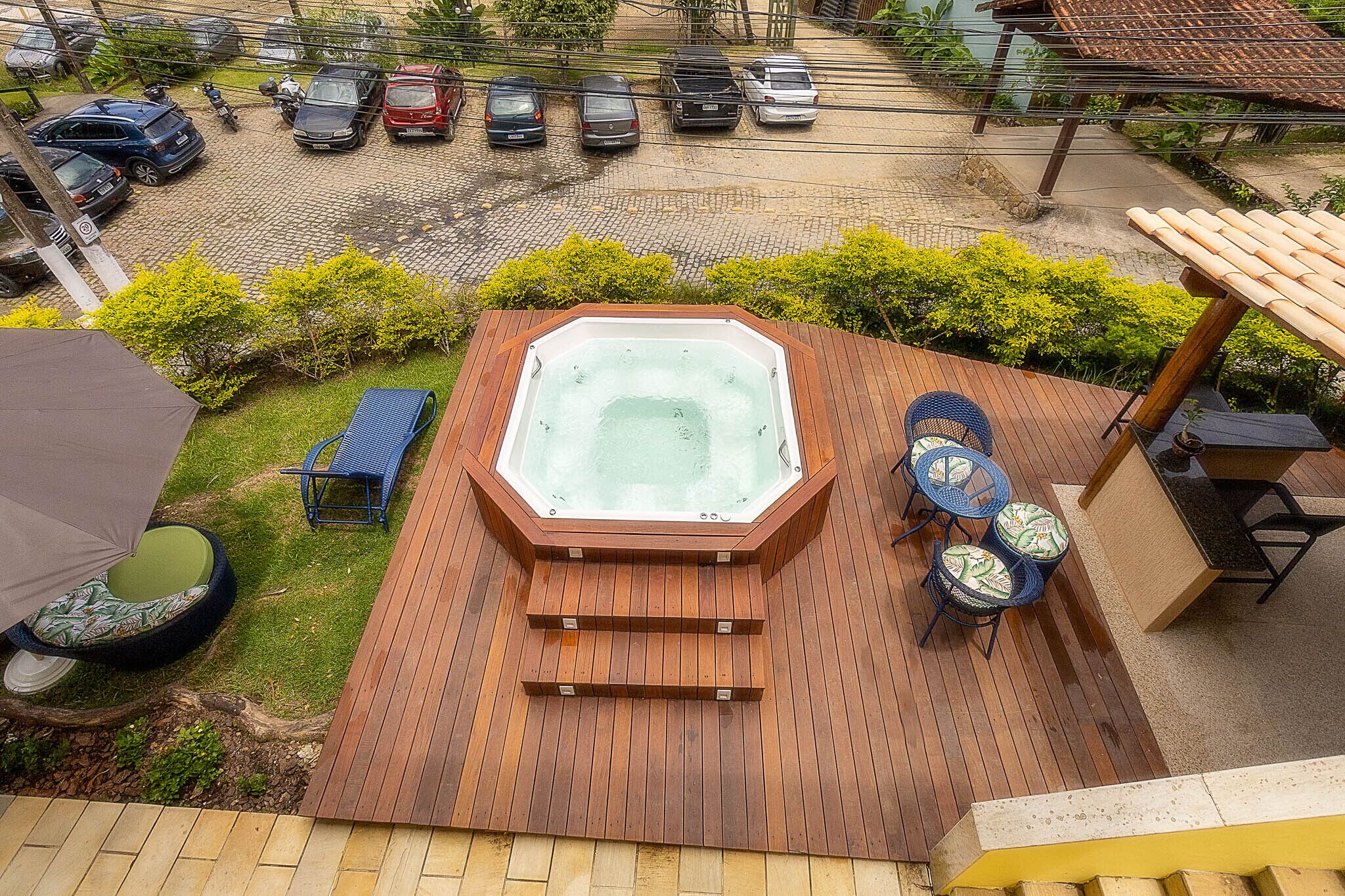 A hot tub on a wooden deck with a view of parked cars and greenery.