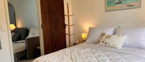 A hotel room with a bed, a desk, a chair, a wooden door, and a painting of a sailboat on the wall.