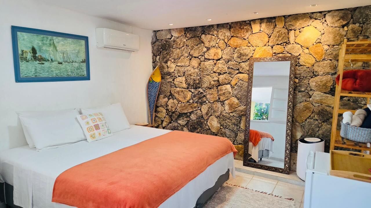 Un dormitorio con pared de piedra, cama con ropa blanca, un cuadro, un espejo y un estante de madera con un cojín rojo.