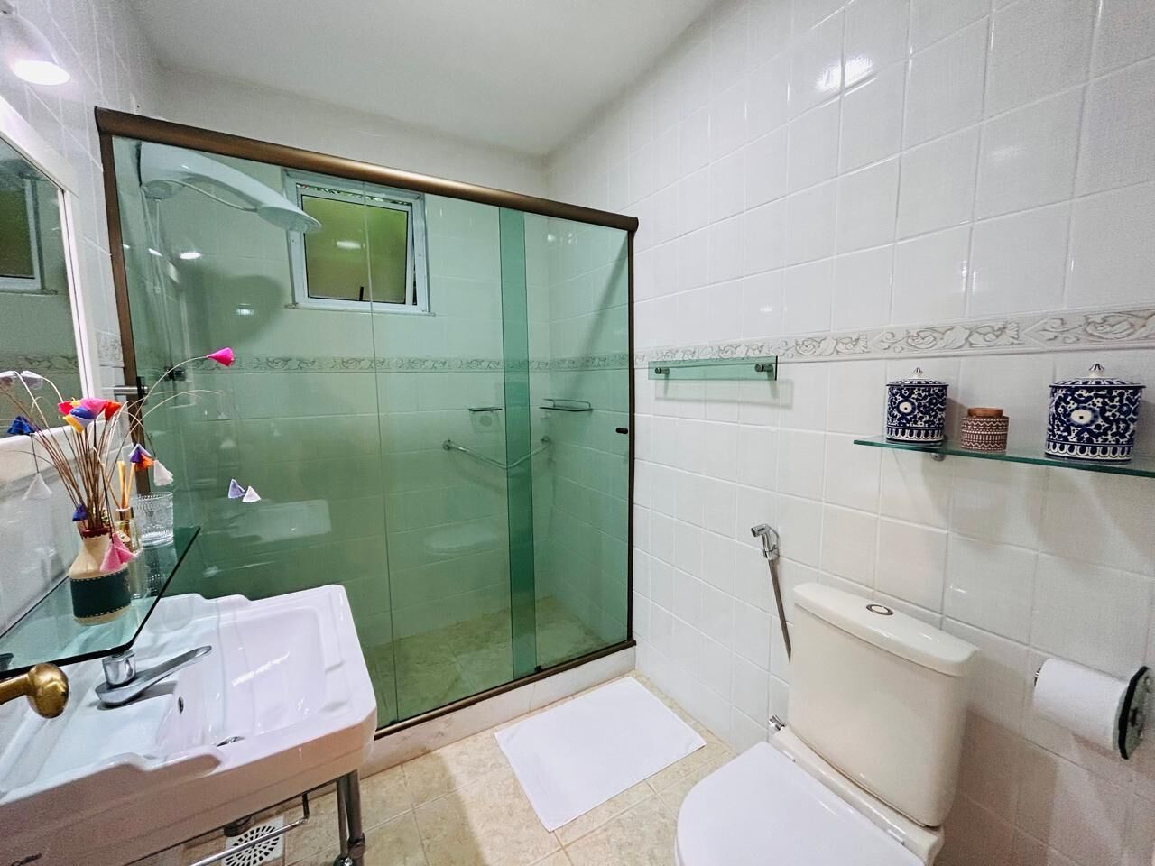 A bathroom with a glass-enclosed shower, a toilet, a sink with a mirror, and a shelf with decorative items.