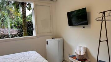 A bedroom with a bed, a small white refrigerator, a TV mounted on the wall, a window with a view of palm trees, and a wooden ladder.