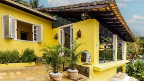 Un edificio amarillo con contraventanas blancas, puerta de cristal y un pequeño balcón con plantas en macetas.