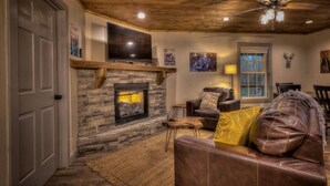 A cozy living room with a stone fireplace, leather furniture, and a ceiling fan. - 5-Star Cabin w/Hot Tub, Game Room, & Resort Perks! (Ellijay)
