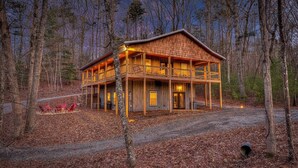 A two-story log cabin with a wrap-around porch, surrounded by a forest. - 5-Star Cabin w/Hot Tub, Game Room, & Resort Perks! (Ellijay)