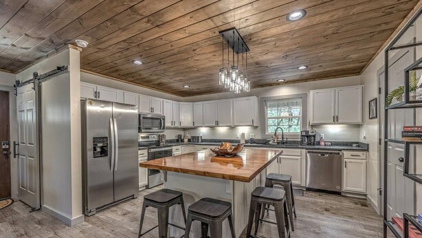 A modern kitchen with a wooden ceiling, stainless steel appliances, a central island with stools, and a large window. - 5-Star Cabin w/Hot Tub, Game Room, & Resort Perks! (Ellijay)