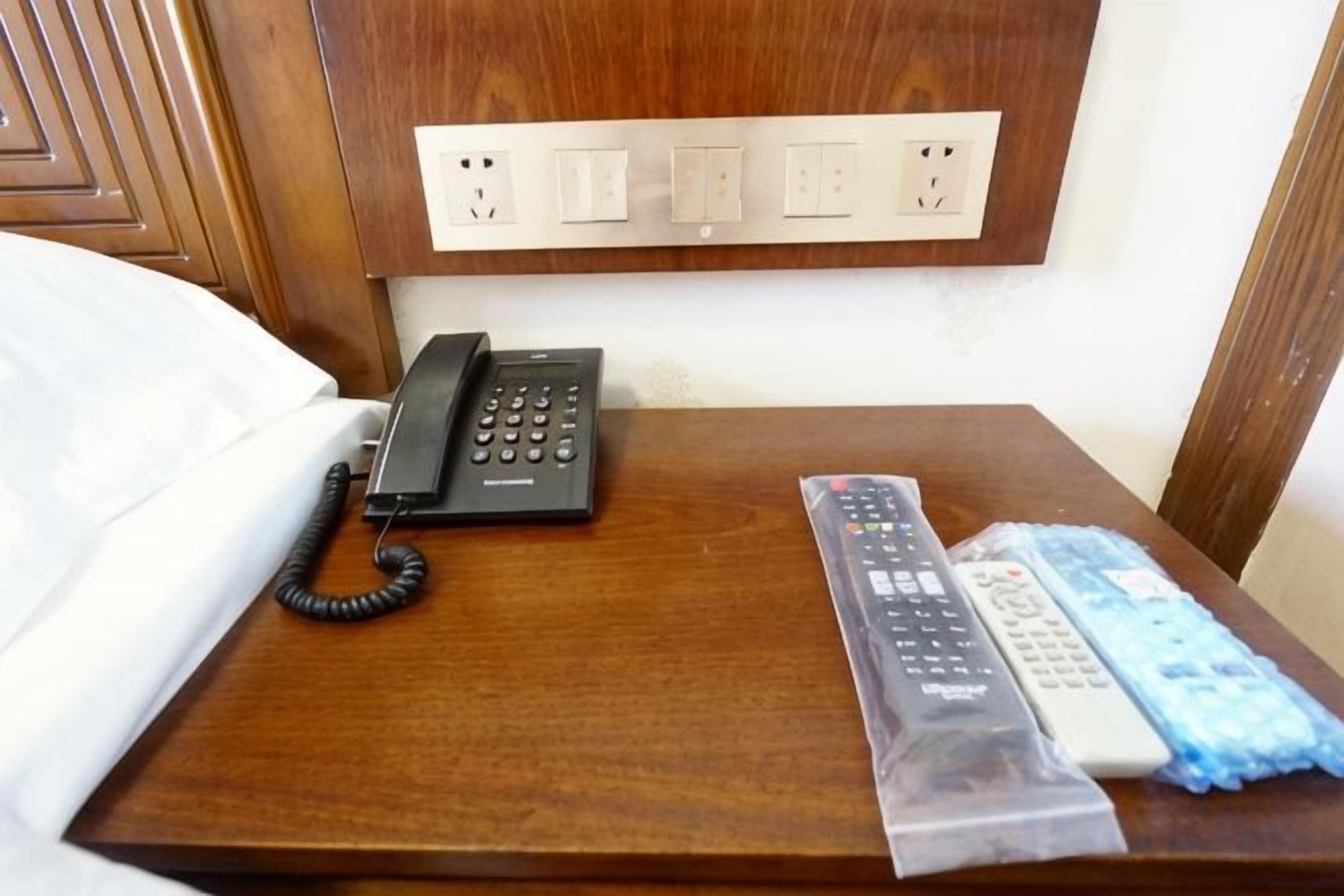 A wooden bedside table with a telephone, a remote control, and a plastic bag containing additional remotes.