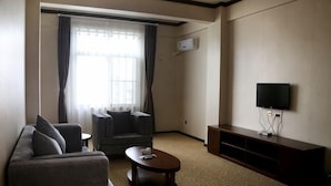 Habitación de hotel con sofá, sillón, mesa de centro, televisor y un ventanal con cortinas.
