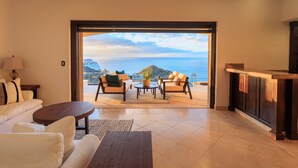 A spacious living area with a sofa, coffee table, and chairs, leading to a balcony with a view of the sea and a distant island. - Casa Vista Tranquila  (Cabo San Lucas)