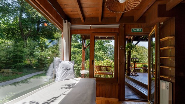 A wooden cabin with a bed, a bench, and a view of a garden and road. - Finca La Peñiga (Mazcuerras)