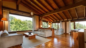 A spacious living room with wooden flooring, a sofa, a coffee table, and a dining area with a table and chairs. Large windows offer a view of greenery outside. - Finca La Peñiga (Mazcuerras)