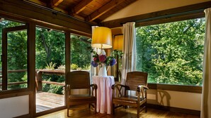 A room with wooden flooring, two wooden chairs, a table with a white tablecloth, a yellow lamp, and a vase with flowers. Large windows offer a view of greenery outside. - Finca La Peñiga (Mazcuerras)