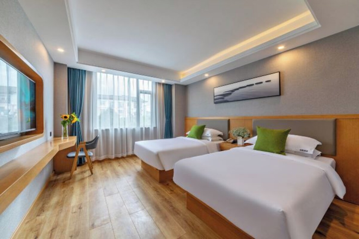 A hotel room with two beds, a desk, a chair, a window with curtains, and a painting on the wall.
