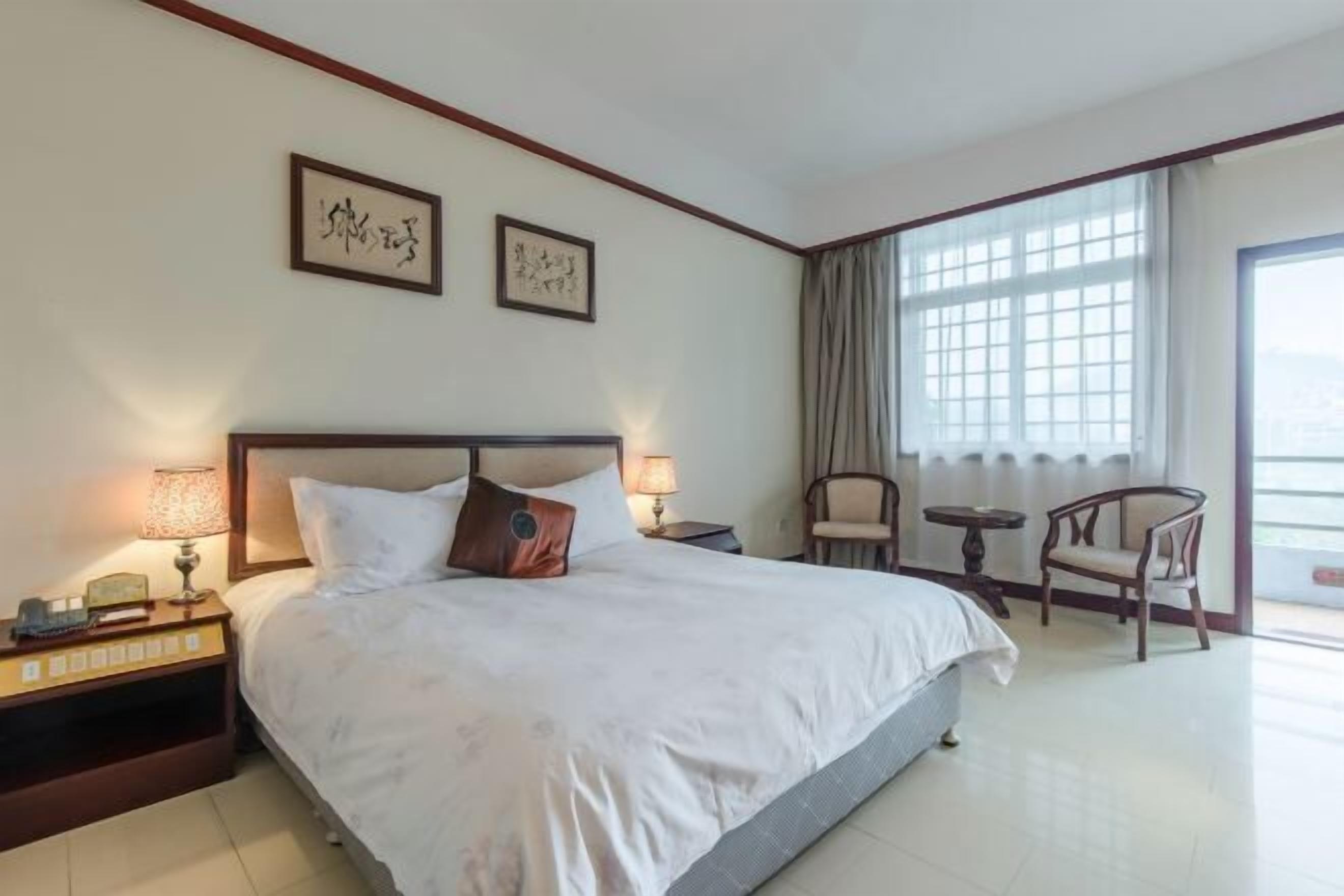 A hotel room with a large bed, two bedside tables with lamps, a chair, a small table, and a large window with curtains.