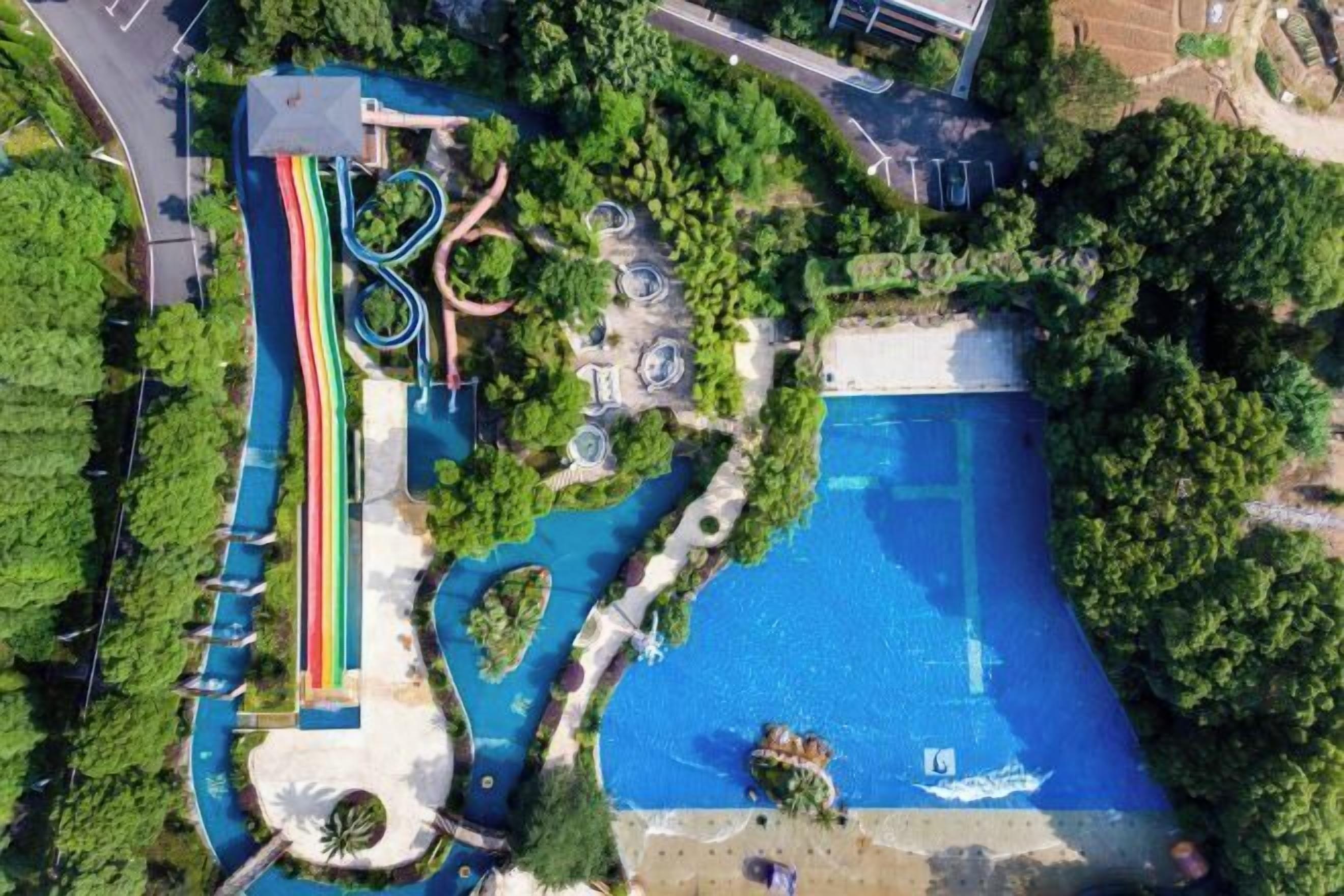 An aerial view of a park with a swimming pool, slides, and walking paths surrounded by greenery.