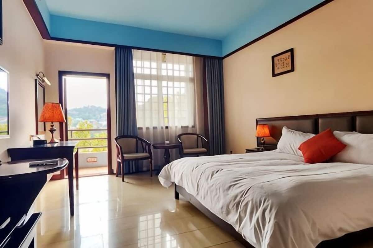 A hotel room with a bed, a desk, a chair, a table, a lamp, and a window with curtains.