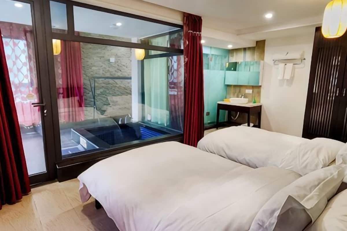 A hotel room with a glass sliding door leading to a balcony, a bed with white bedding, a small bedside table, and a wall-mounted sink.