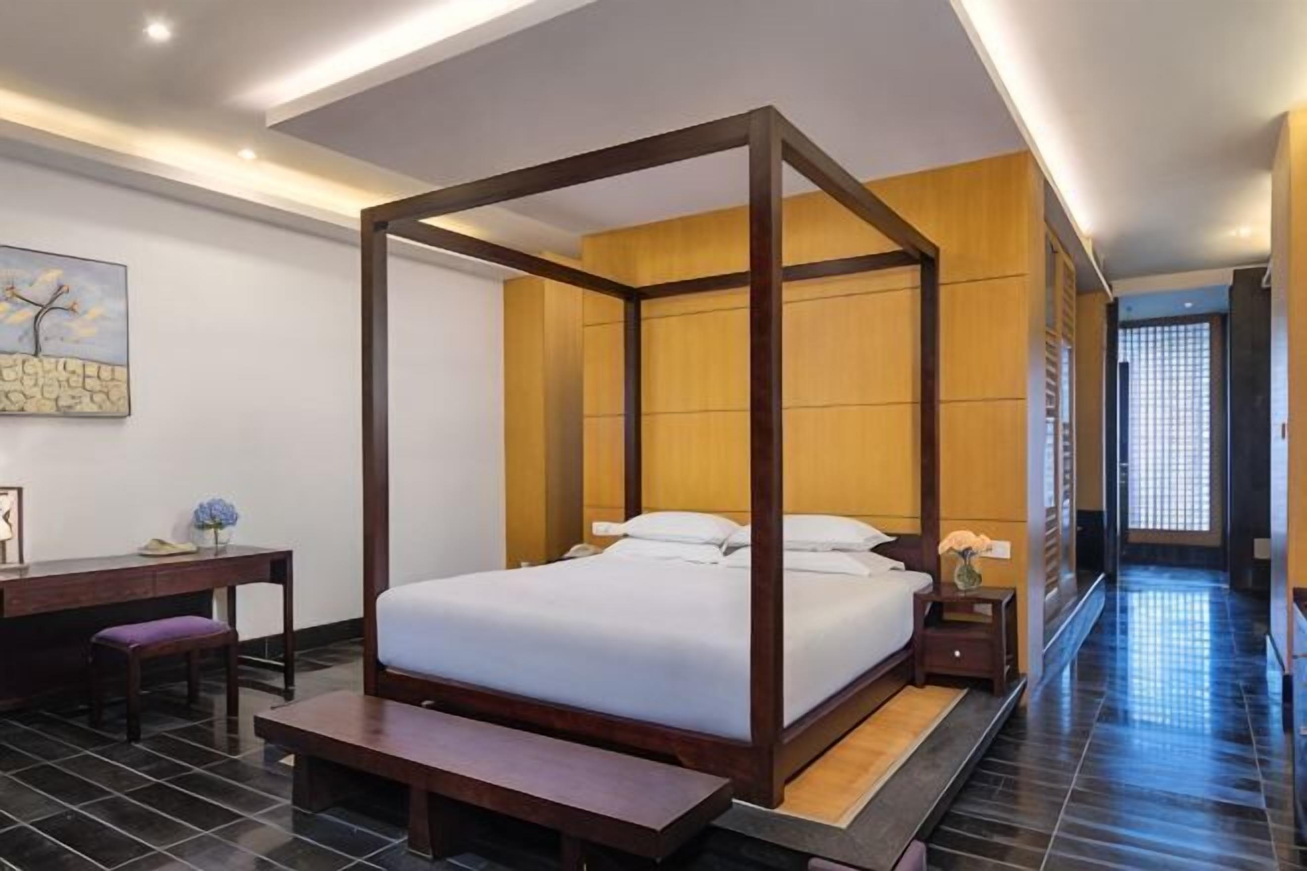 A four-poster bed with a wooden frame, a desk with a chair, and a bench in a hotel room with tiled flooring.