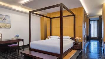 A four-poster bed with a wooden frame, a desk with a chair, and a bench in a hotel room with tiled flooring.
