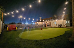 Zona exterior bien iluminada con un putting green, un bolso de golf rojo y amarillo, y un edificio con un porche cubierto.