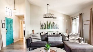 Un salon moderne avec un canapé d’angle gris, une table basse ronde agrémentée d’une plante et une grande fenêtre avec des rideaux.