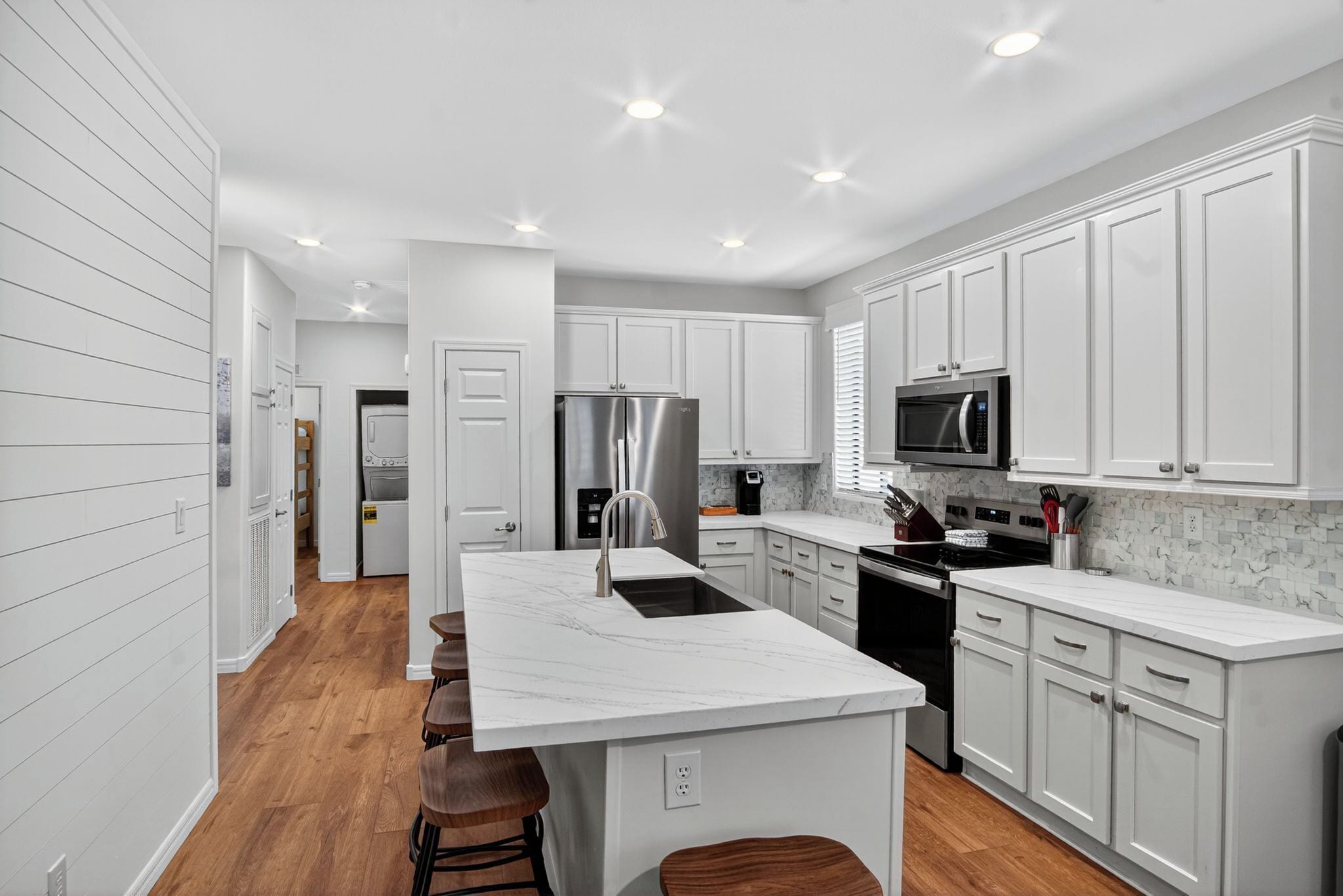 A modern kitchen with white cabinetry, a marble island, stainless steel appliances, and a wooden bar stool seating area.