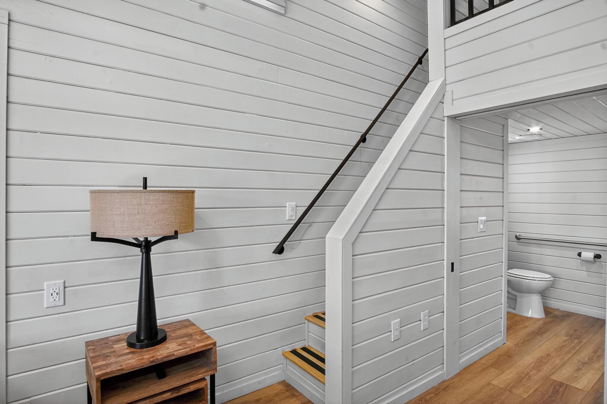 A staircase leading to a second floor, a wooden nightstand with a lamp, and a toilet in a room with white paneled walls.