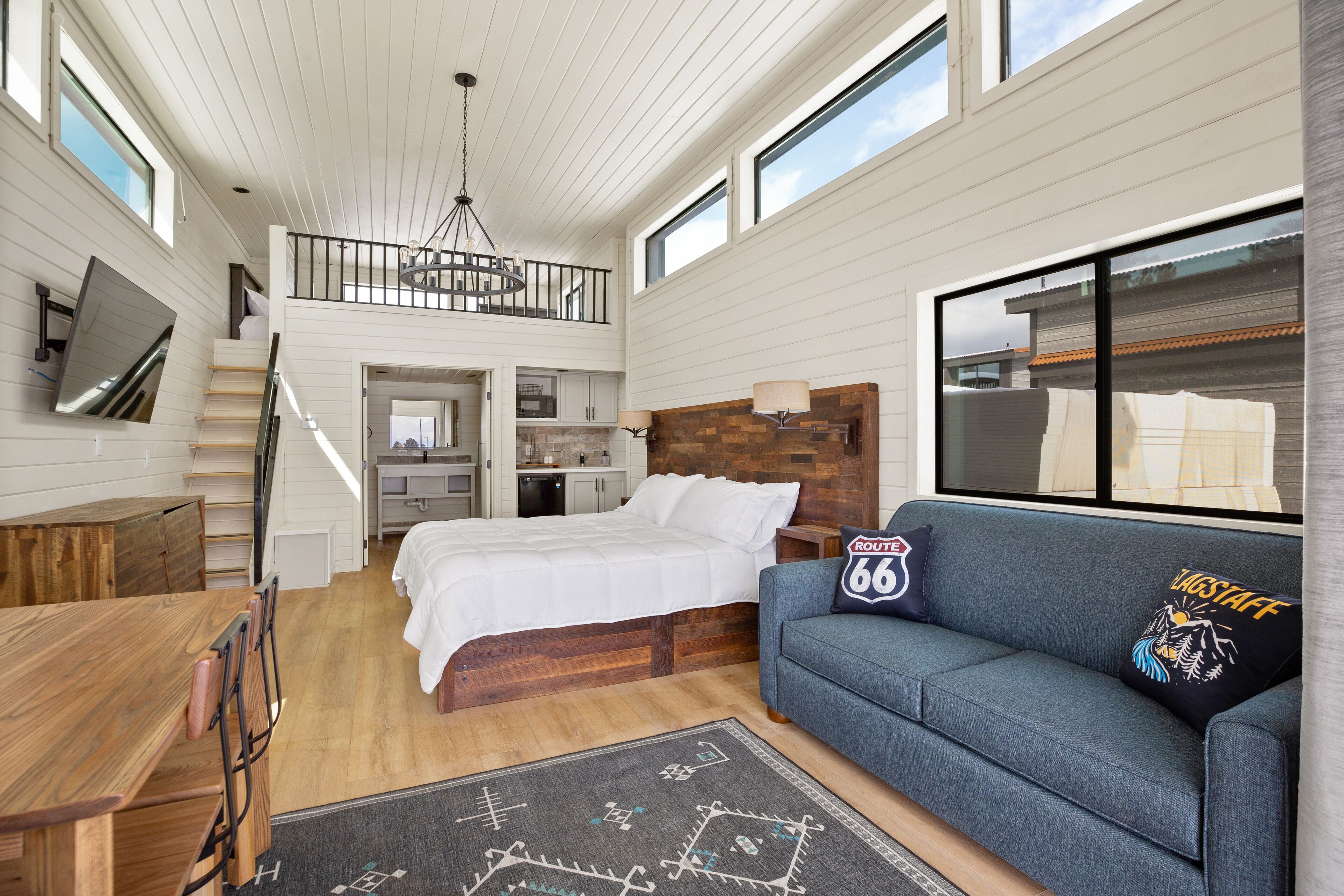 A spacious bedroom with a large bed, a sofa, a wooden dining table, and a staircase leading to an upper level.