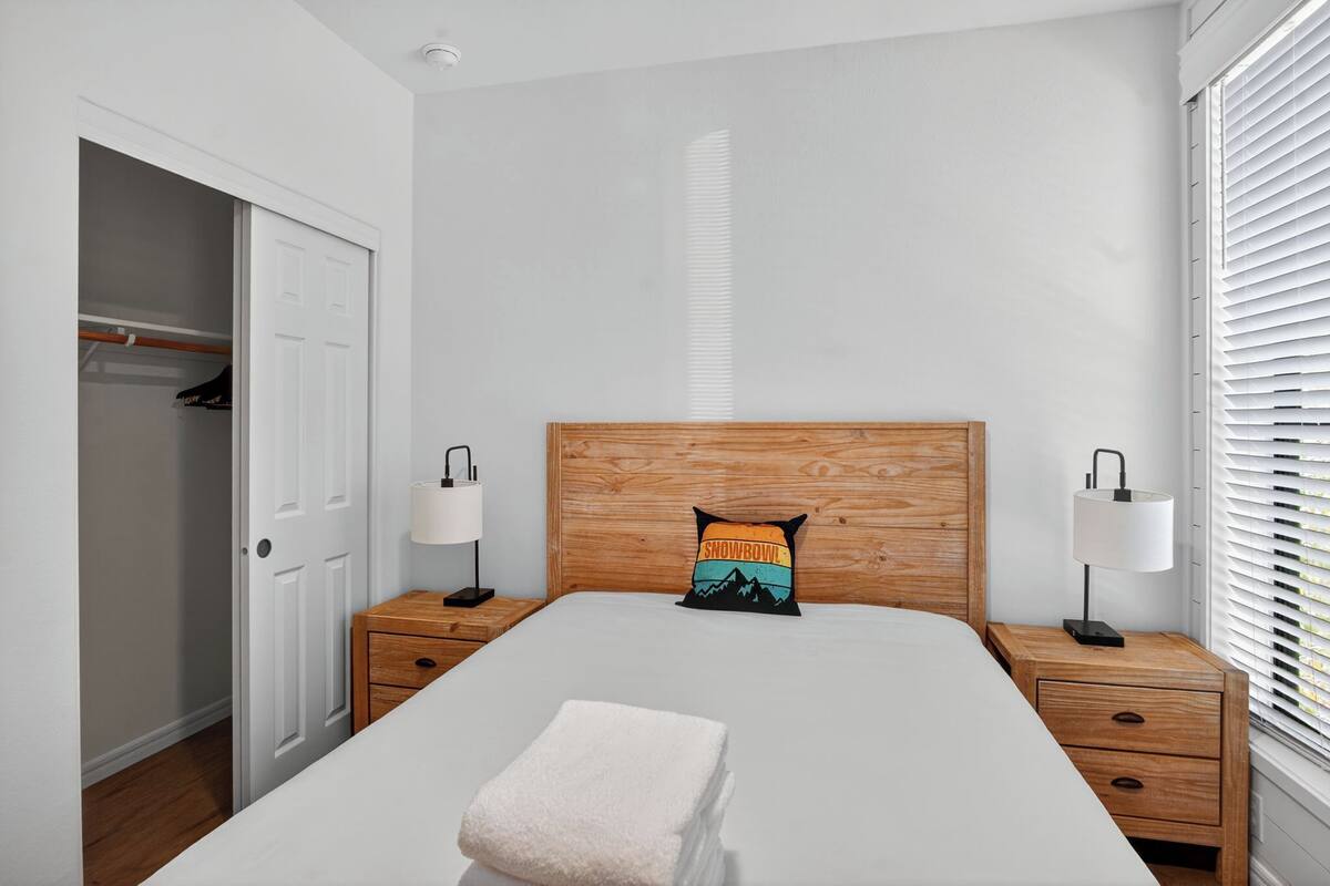 Une chambre à coucher avec une tête de lit en bois, une table de chevet avec une lampe, un placard et une fenêtre avec des stores.