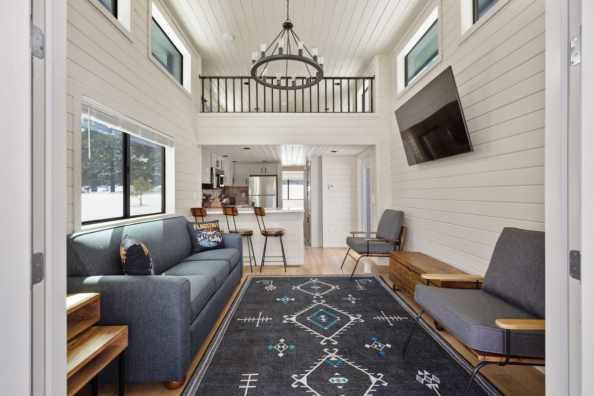 Un vaste espace de vie agrémenté d’un canapé gris, d’un banc en bois et d’un tapis à motifs. On aperçoit à l’arrière-plan un coin repas avec une table et des chaises.