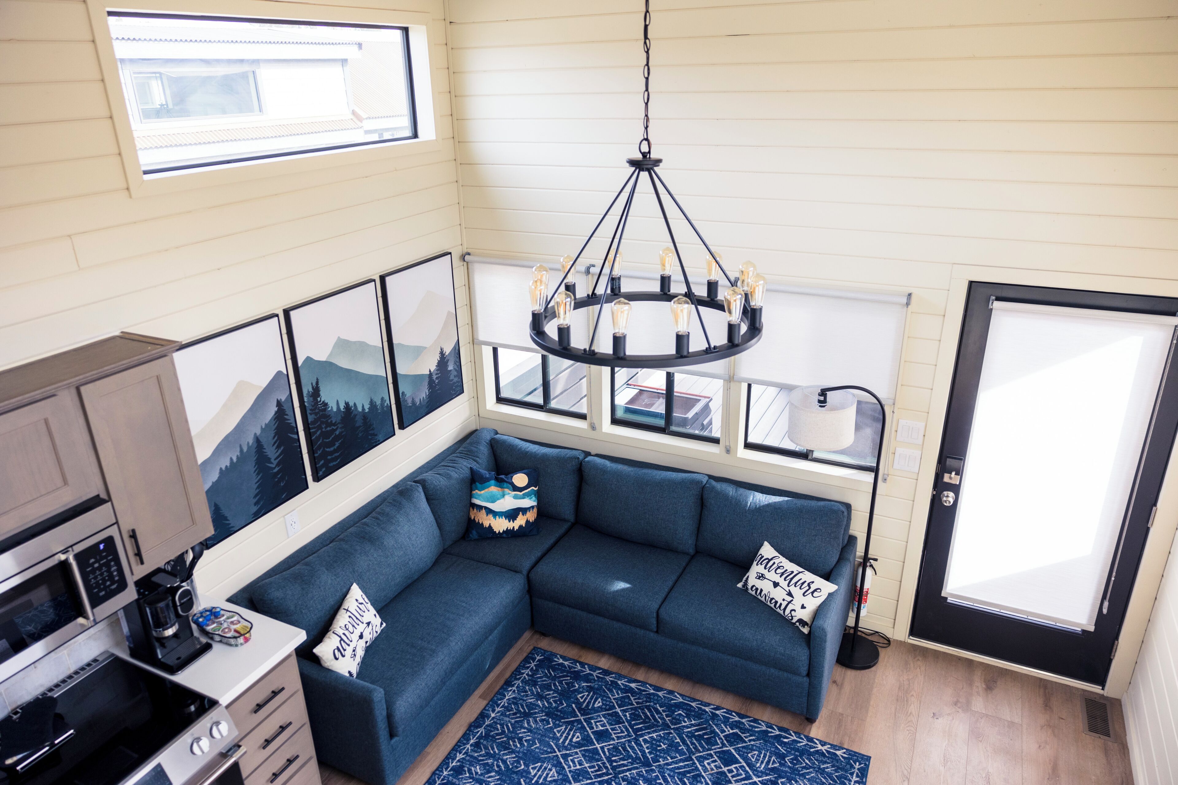 A cozy living area with a sectional sofa, a chandelier, a kitchen with a microwave, and framed artwork on the walls.