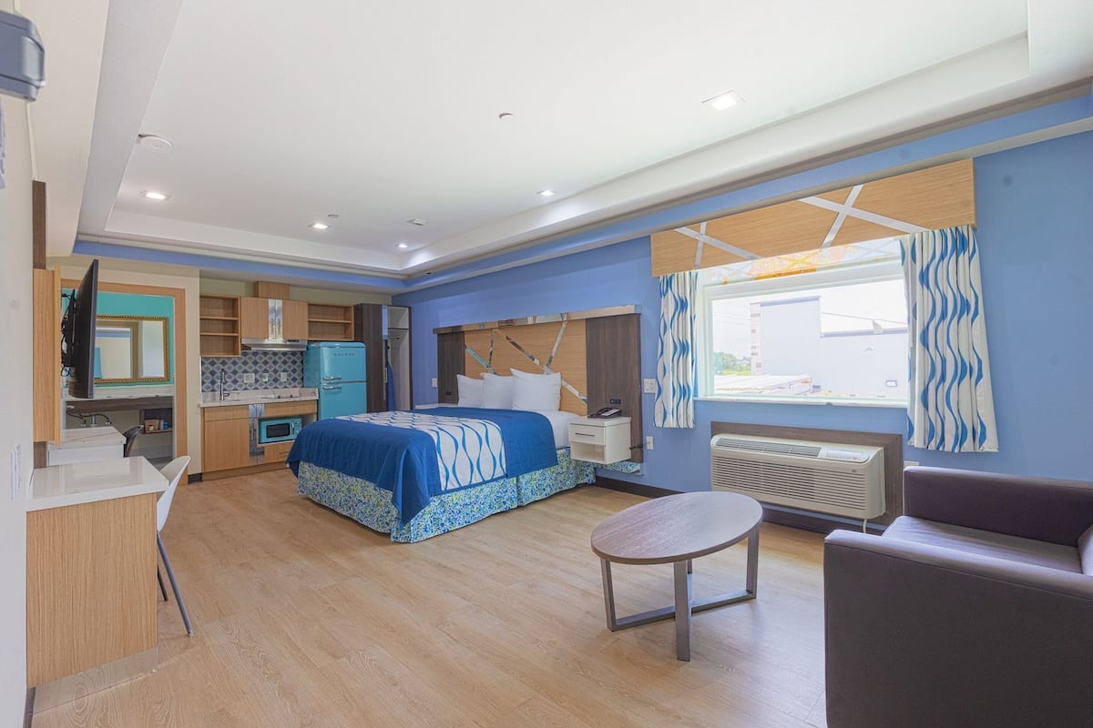 Una camera d'albergo compatta con un letto, un piccolo tavolo, una divisa, angolo cottura e una finestra con tende.