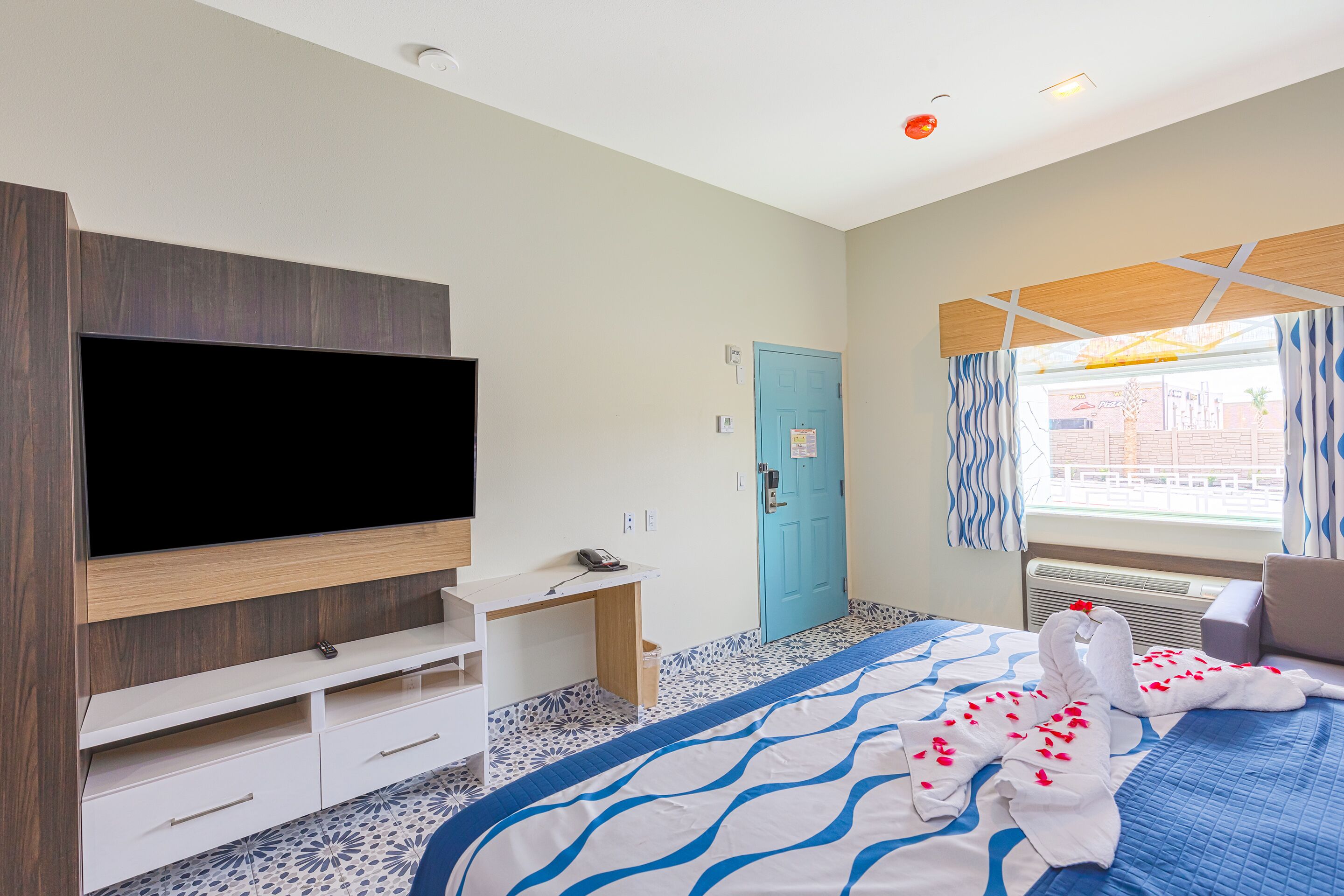 A hotel room with a large flat-screen TV, a desk, a bed with a blue and white patterned bedspread, and a window with blue curtains.