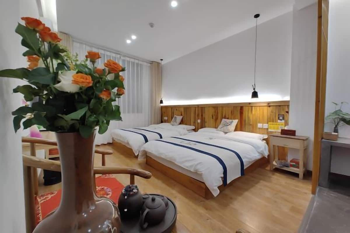 A hotel room with two beds, a wooden headboard, a bedside table with a lamp, a vase with flowers, and a teapot on the floor.