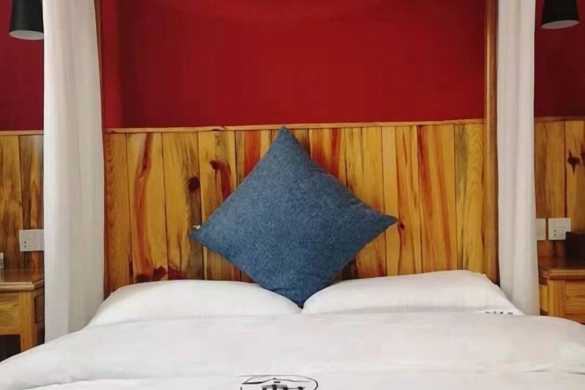 A bed with a blue pillow, white bedding, and a wooden headboard with a red wall behind it.