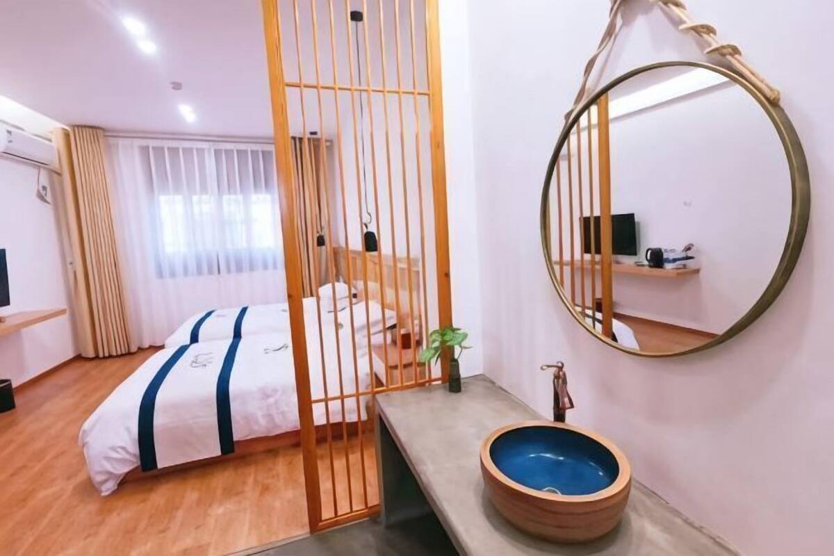 A hotel room with a bed, a wooden partition, a round mirror, a sink with a blue basin, and a window with curtains.