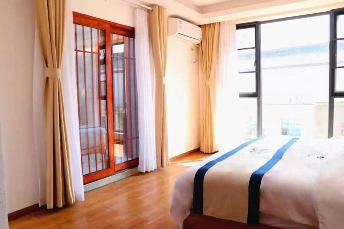 A hotel room with a bed, curtains, an air conditioning unit, and a view through a window.
