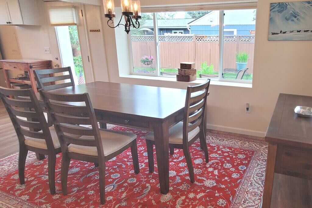 A dining area with a wooden table and chairs, a red patterned rug, and a window with a view of a backyard.
