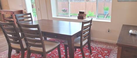 A dining area with a wooden table and chairs, a red patterned rug, and a window with a view of a backyard.