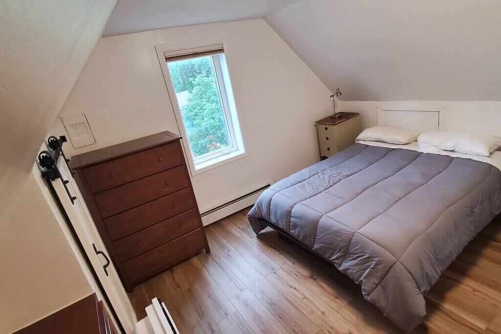 A bedroom with a sloped ceiling, a window, a wooden dresser, a bedside table, and a bed with a gray comforter.