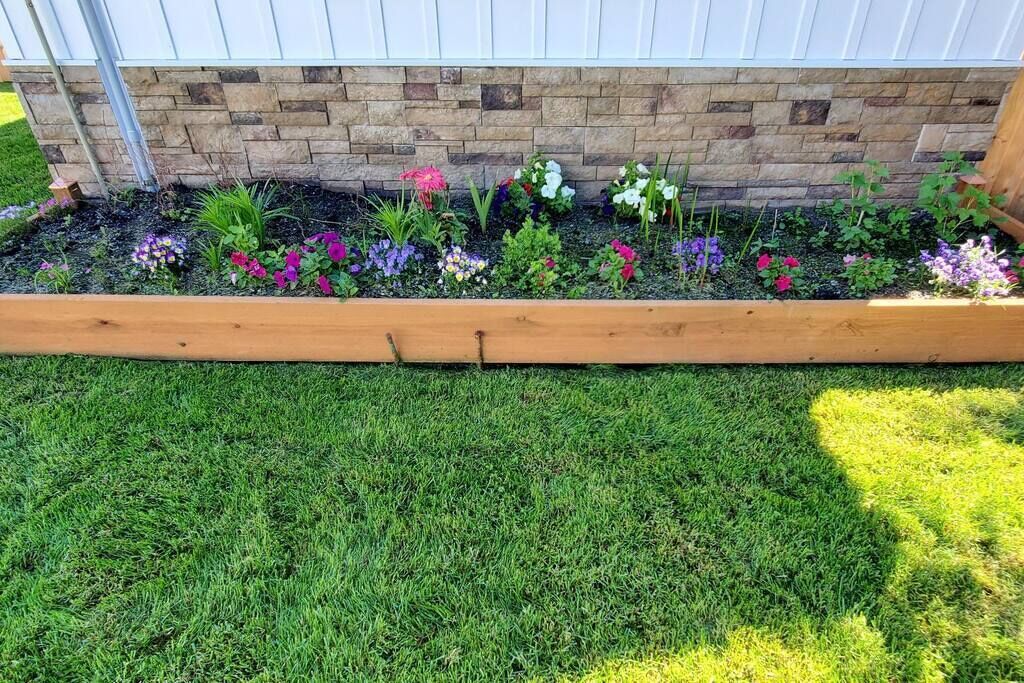 Un jardín elevado con una variedad de flores y plantas coloridas, un borde de madera y un muro de ladrillo al fondo.