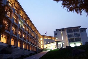 A multi-story hotel with a central courtyard, a flagpole, and a well-lit entrance. - Jinshuiwan Holiday Resort (Shucheng)